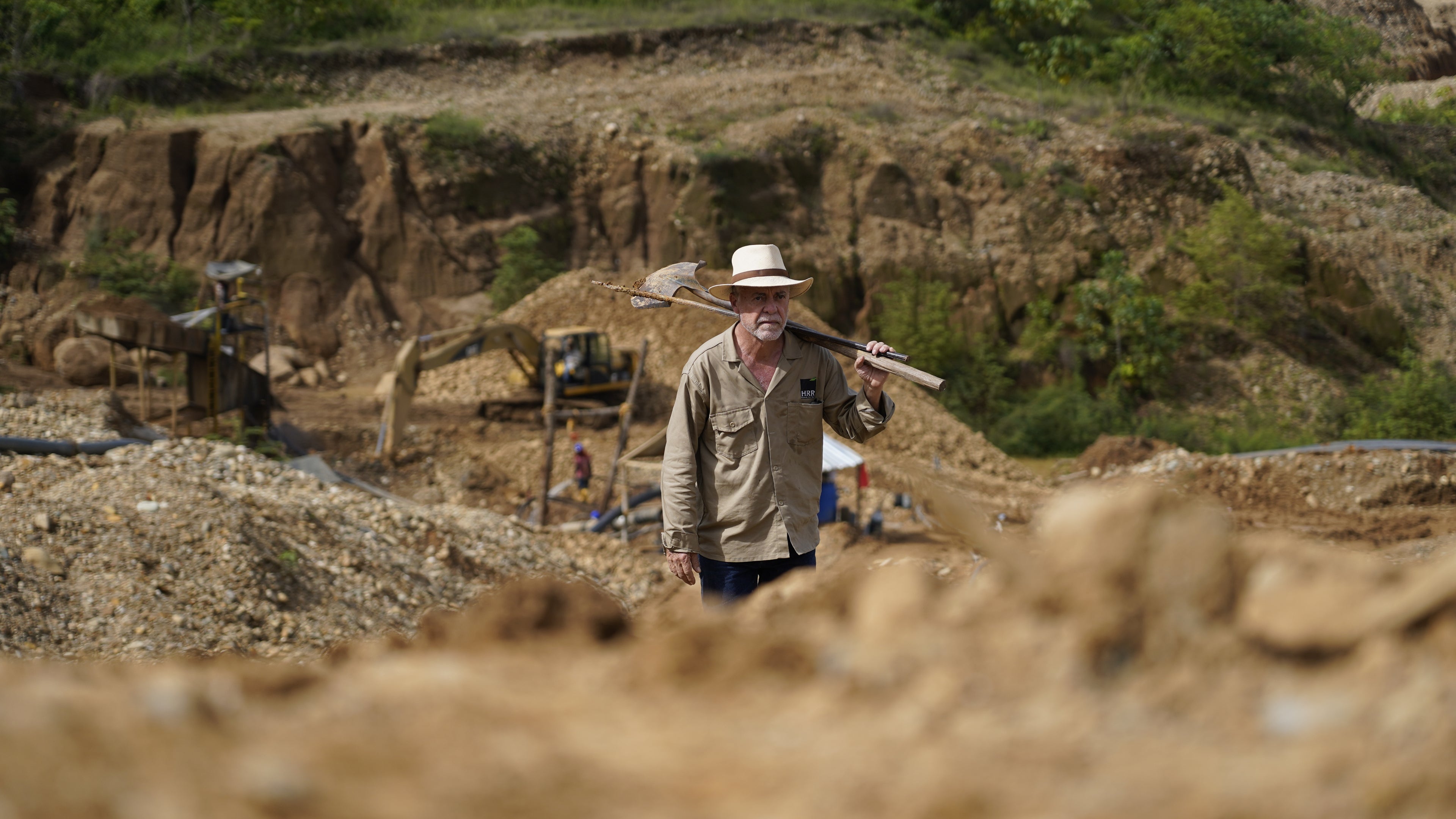 Man with a pickaxe in an excavation site with machinery and workers in the background.